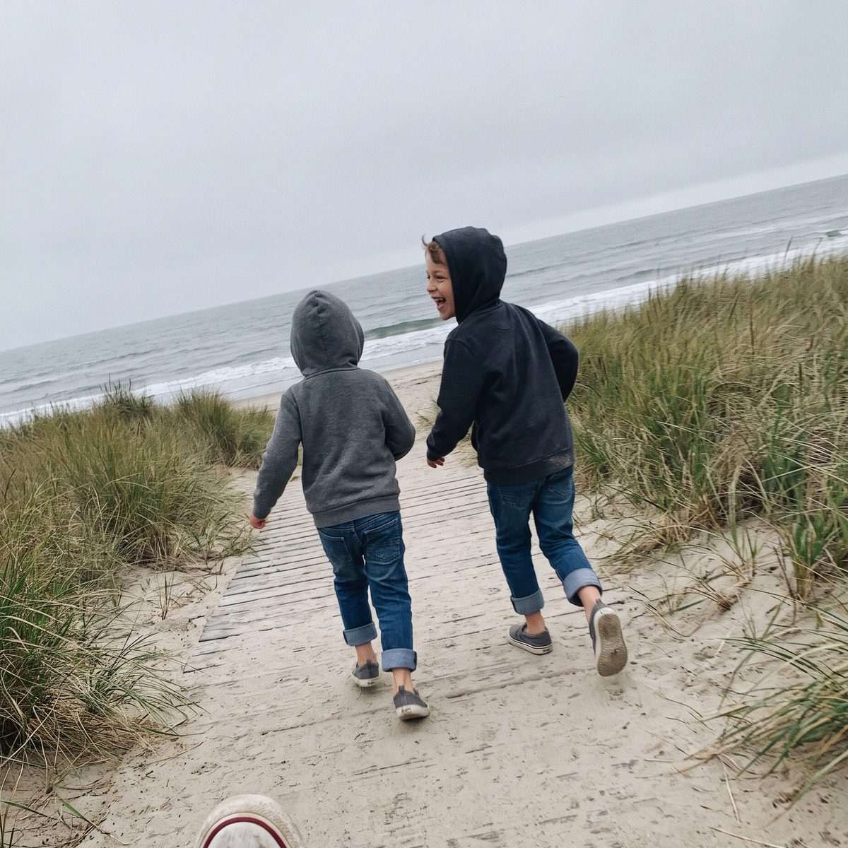 Two small boys running down a sandy boardwalk toward the sea